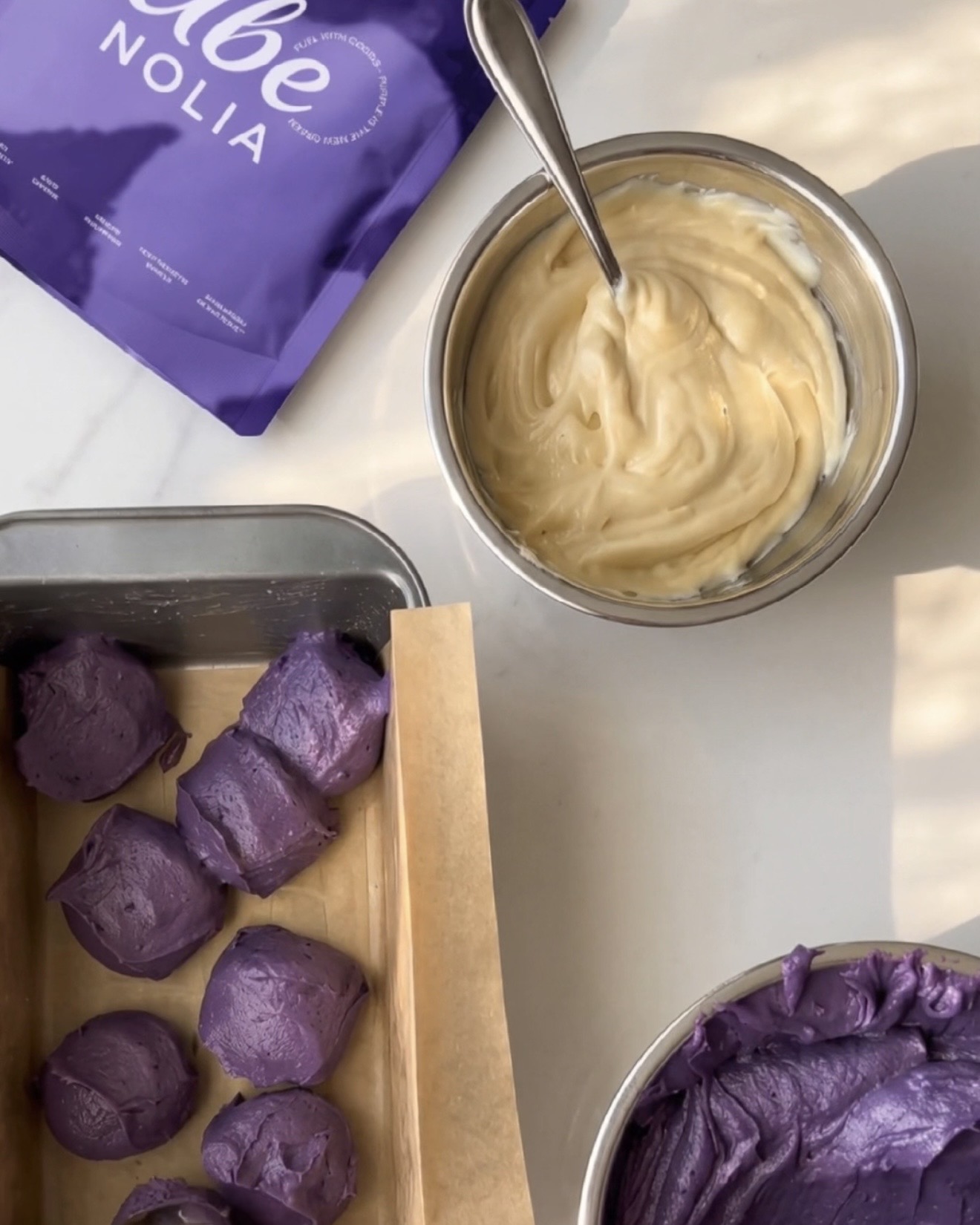 Fertige selbstgemachte Ube Halaya, Vanille-Teig und Schöpflöffel lila Ube-Teig in einer Kastenform neben einem Beutel UBE NOLIA Ube-Pulver